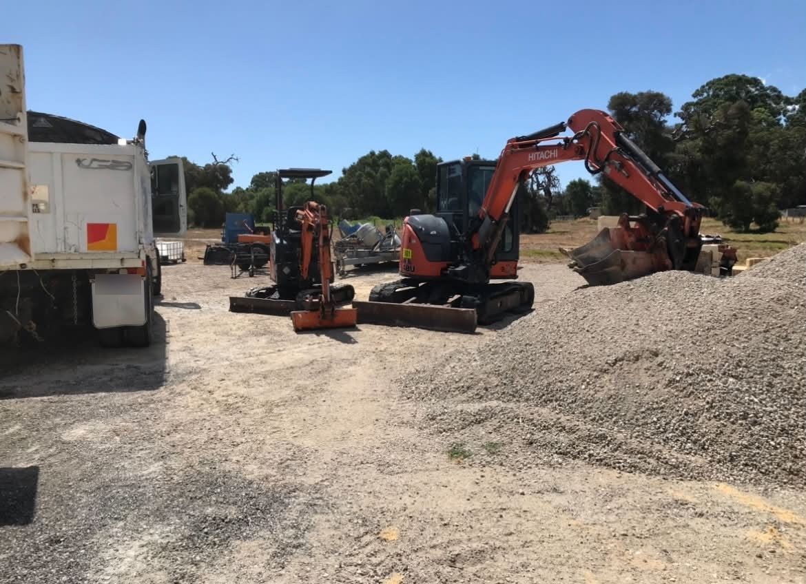 Two Hitachi excavators and a tipper truck beside gravel stockpiles in the yard