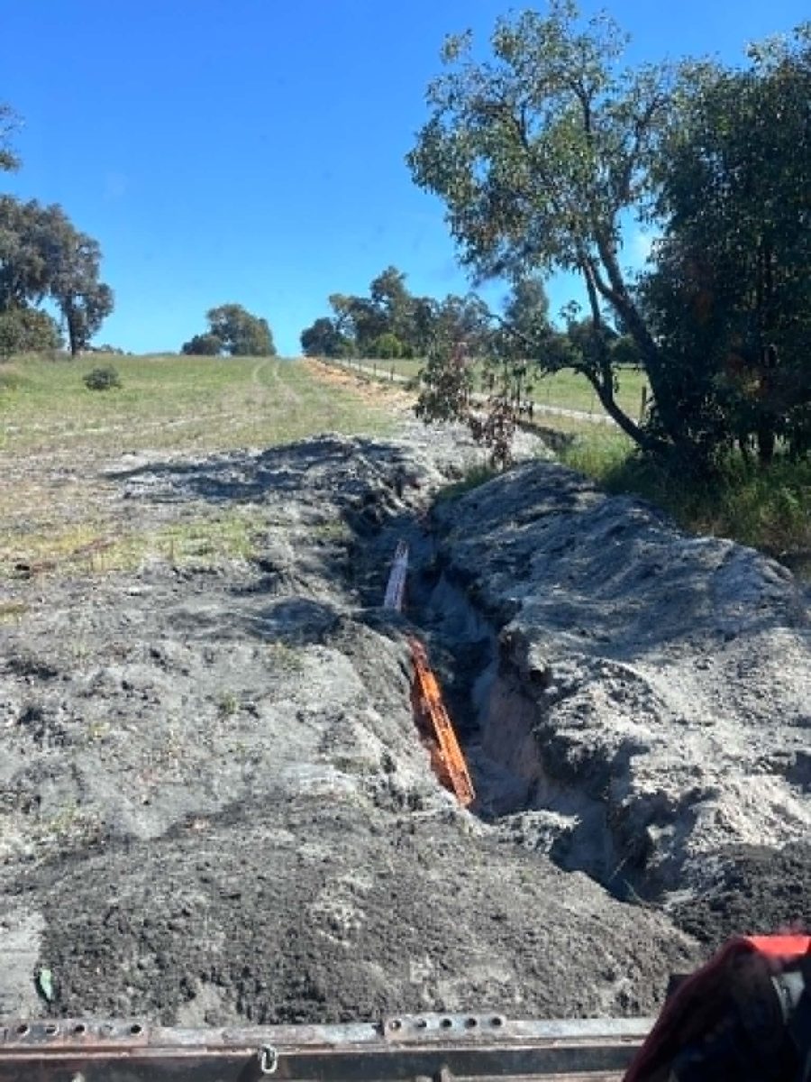 An open trench across a sandy paddock with orange electrical conduit laid in the bottom